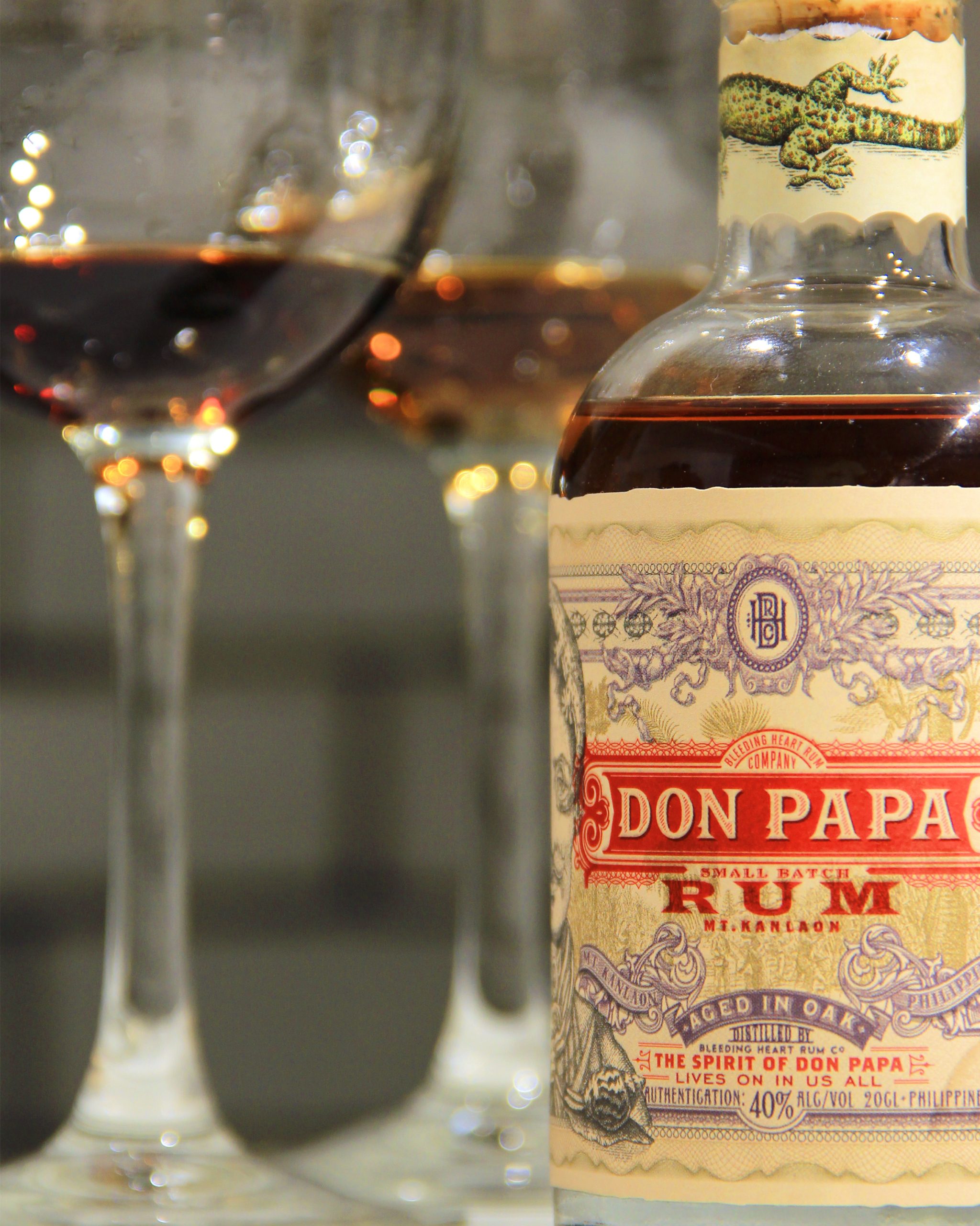 Closeup shot of a bottle of Don Papa Rum, with two wine glasses blurred in front of a grey brick backdrop.