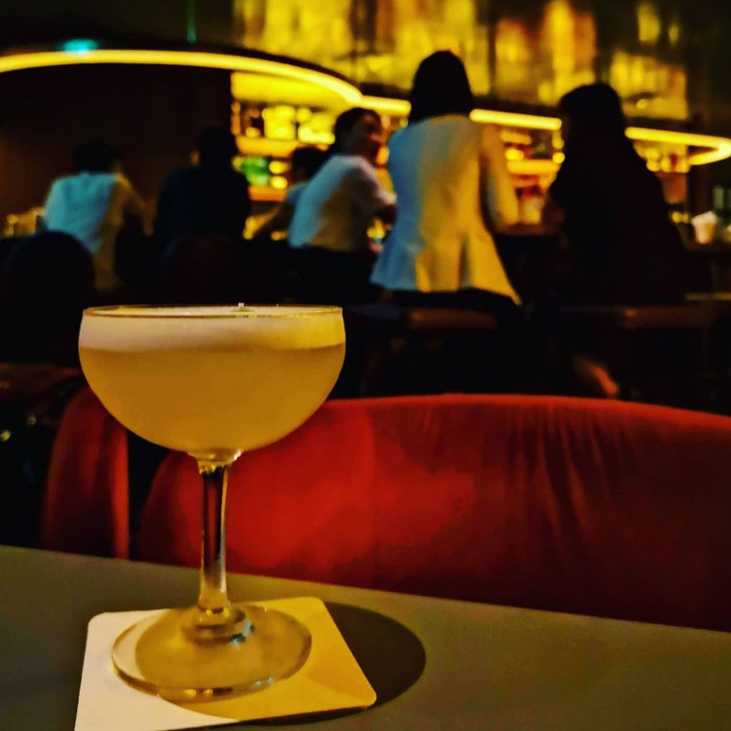 A cocktail glass filled with Jigger & Pony's alcohol-free cocktail, Apple Elderflower. It is placed on top of a white square coaster with rounded edges. Behind the glass, patrons of the bar can be seen talking animatedly among one another. The scene is illuminated by yellowish-orange lights.