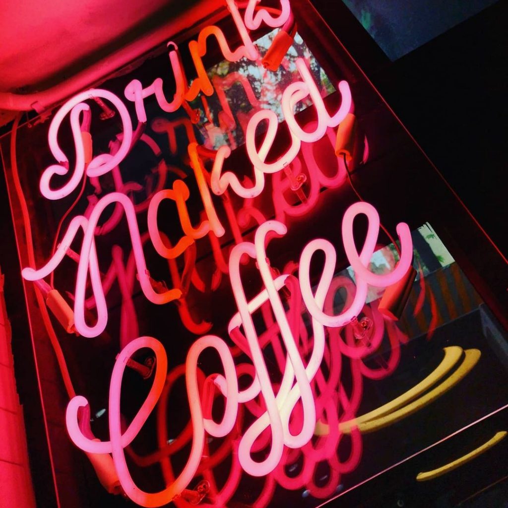 A bright pink neon sign with the cursive words, "Drink Naked Coffee."