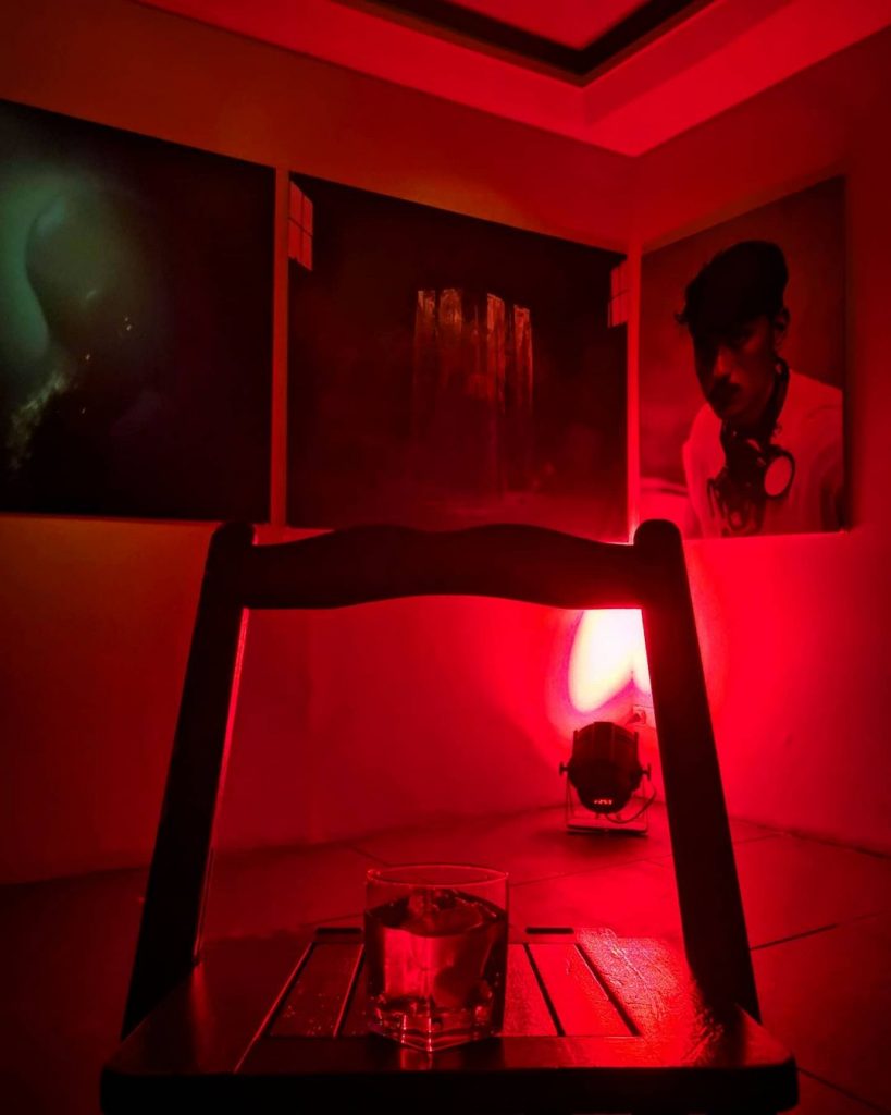 A glass of negroni sits on top of a wooden table in the middle of a room fully-saturated by red light. The walls of the room are adorned with three paintings. Welcome to Limbo.