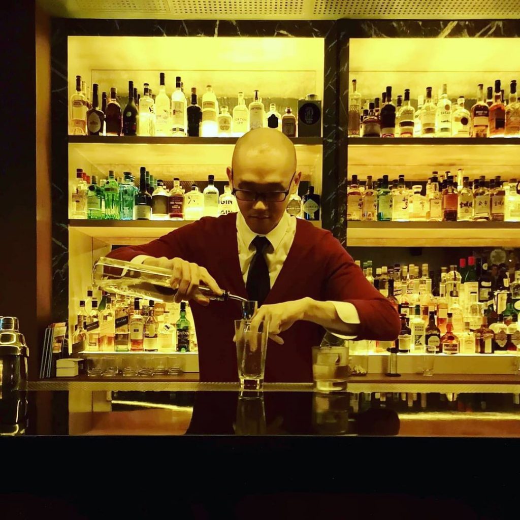 The bartender of Jigger & Pony makes a drink. Behind him are several rows of bottles neatly arranged on shelves behind the bar.