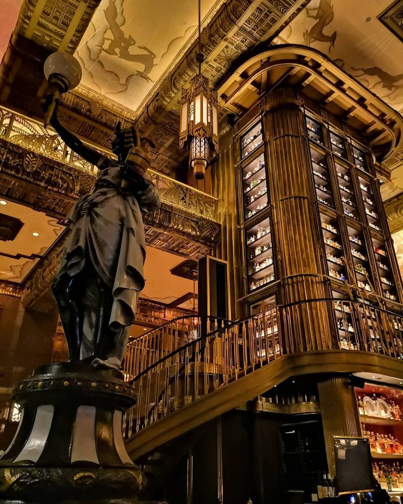 A shot of Atlas's massive ornate pillar, taken from a low vantage point. The base of this pillar serves as the bar itself, which can be barely seen below the image. All sides of the pillar are lined with shelves from top to bottom, each of them fully-lined with bottles of gin. To the left of the photo, a Greco-Roman statue stands proudly akin to the Statue of Liberty, holding a torch on the left hand and a shaker on the left. The art-deco-inspired designs on the ceiling can be seen.