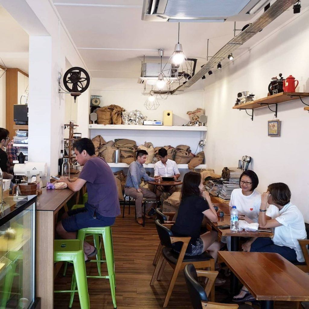 A photo of Tiong Hoe Specialty Coffee's interior.  The chairs, tables, floor, and bar are made predominantly of wood, with the exception of green stools serving as bar chairs. Several brown burlap sacks sit on top of three tiers of shelves occupying the entirety of the background wall. The lone barista behind the bar serves six customers: one sitting in front of the bar, three by the long table to the right of the photo, and two by the round table in front of the burlap sacks.
