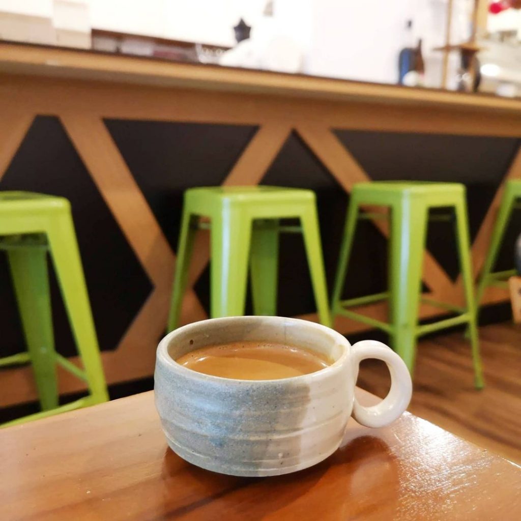 A ceramic espresso cup filled with espresso occupies the foreground, sitting on the corner of a brown wooden table. The background is occupied by the bar of Tiong Hoe Specialty Coffee, lined by tall green stools.