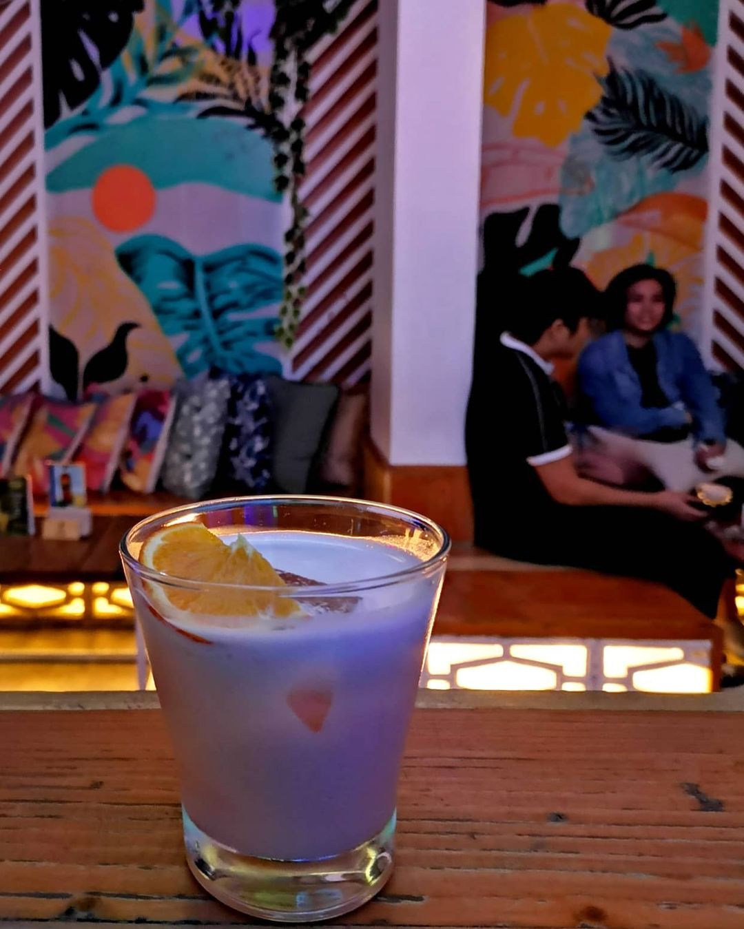 A glass of Tortuga's Coco Crush signature cocktail, a drink made of coconut rum, Amarula, and Bianco. It is garnished with fresh lime and an orange slice. The glass is set on top of an orange-painted bamboo platform on the foreground, with Tortuga's beach-themed interior serving as the background.