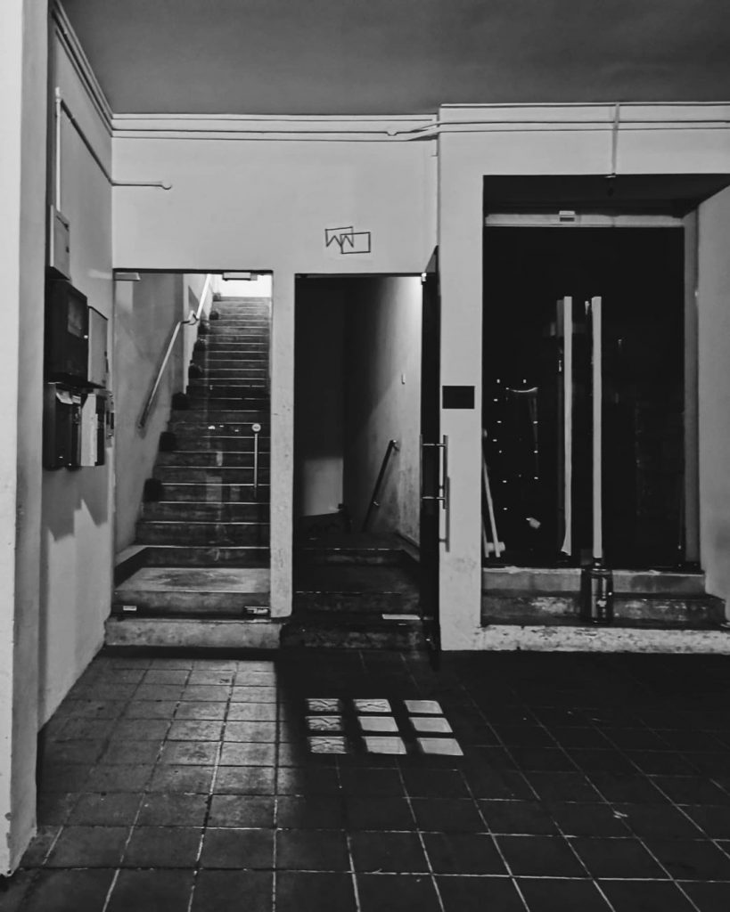 An exterior shot of the entrance to Operation Dagger, in grayscale. The passageway to the bar is an open door, which is flanked by a staircase on the left side of the photo and a closed set of doors on the right. The door to Operation Dagger has its logo etched on the wall above the doorframe. The passageway has a staircase leading down towards an area which cannot be seen on the photo.