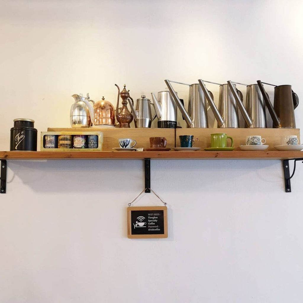 A shot of a wooden shelf hanging on Tiong Hoe Specialty Coffee's wall. Sitting on top of the shelf are various knick-knacks: five special coffee jars, nine fancy-looking kettles, and six coffee cups (on saucers) of varying designs. Hanging below the shelf is a framed signage bearing Tiong Hoe Specialty Coffee's wifi SSID and password.