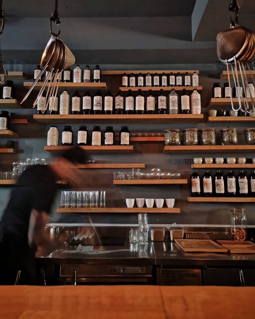 A closeup shot of the gray wall behind Operation Dagger's bar. At least fifteen horizontal wooden shelves are installed on the wall, with neatly-arranged bottles, jars, and glass containers sitting on top of each one. Below these shelves are the metal counters where the bartender does his work. On the photo, he appears to be crushing ice. A portion of the bar can be seen in the foreground, as well as sets of strainers hanging down from the top of the photo.