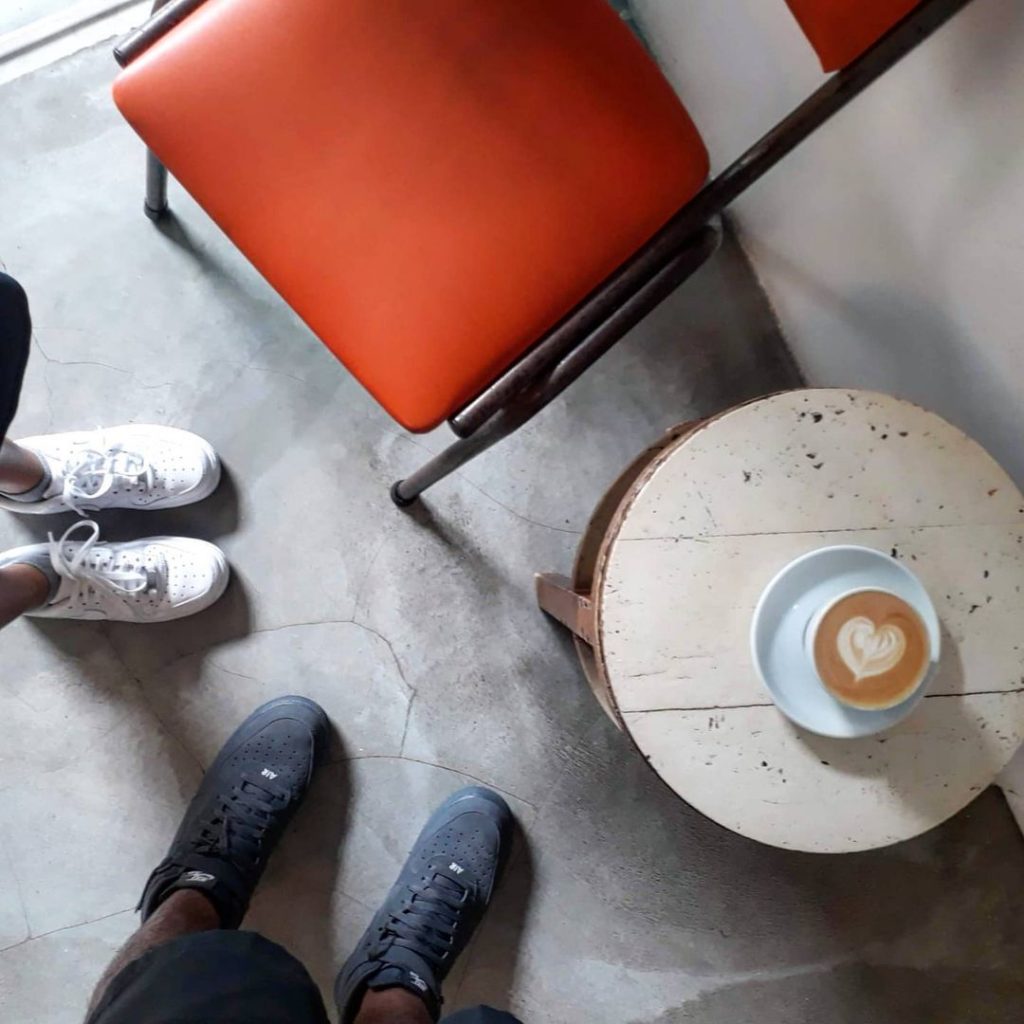 An overhead shot of Nylon Coffee's interior, showing its concrete floor and white wall. The feet of two people, possibly a couple, stand beside one another, both facing a red chair and a small, round wooden coffee table. On top of the table is a cup of coffee with a heart-shaped latte art.