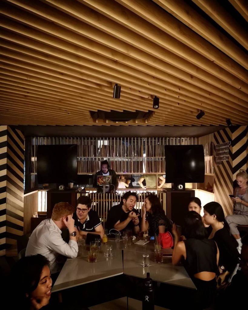 A shot of OTO's interior. Nine people are sitting around a polygonal table, chatting animatedly among one another as a DJ plays music behind the bar in the background. The brown and black aesthetic of the architecture is dominated by straight lines of various orientations, but mostly parallel with one another.