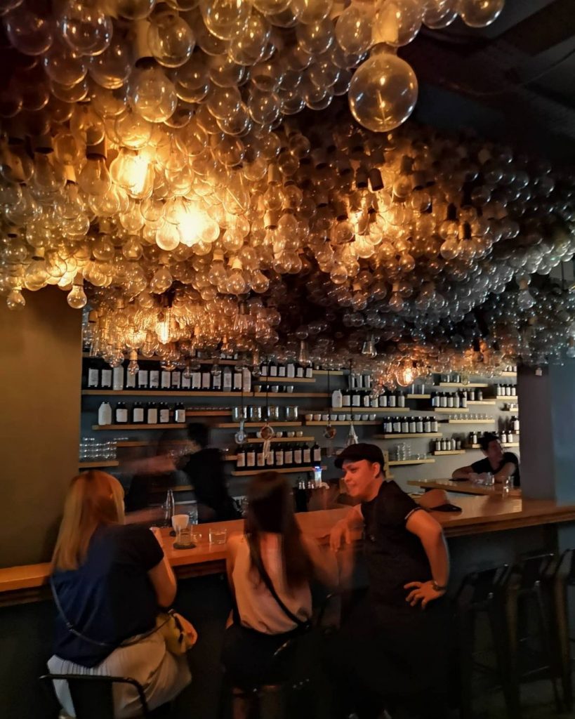 A shot of Operation Dagger's interior. In the foreground, three people can be seen sitting in front of the wooden bar, with another person sitting on the far right end. The bartender is busy making a drink behind the bar, facing a gray wall that's covered with several wooden shelves full of neatly-arranged bottles. The scene is illuminated by several lightbulbs hanging down the ceiling, which cannot be seen due to the sheer number of bulbs covering it.