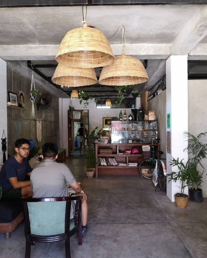 A shot of the predominantly-grey interior of Masa Cafe. One wall of the cafe (left side of photo) is made with the same smooth concrete as the cafe floor. The wooden bar can be seen near the center of the photo. A bicycle is parked beside it. Hanging plants and basket-wrapped lamps hang from the ceiling. Two people having a conversation serves as the foreground of the image.